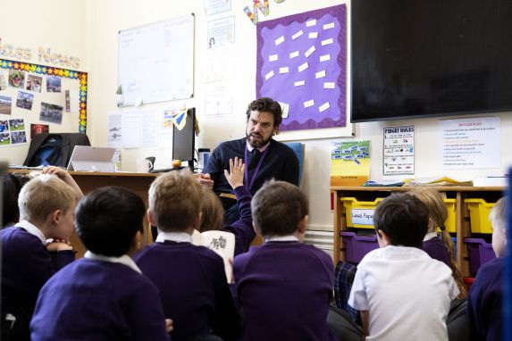 Male prep school teacher with group of pupils. One pupil with hand up.