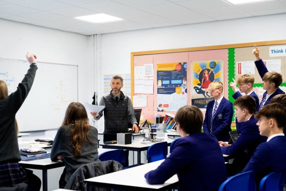 Teacher surrounded by school pupils in classroom with hands up