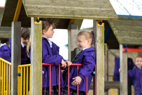 two pre prep girls in purple coats talking on wooden outdoor play equipment